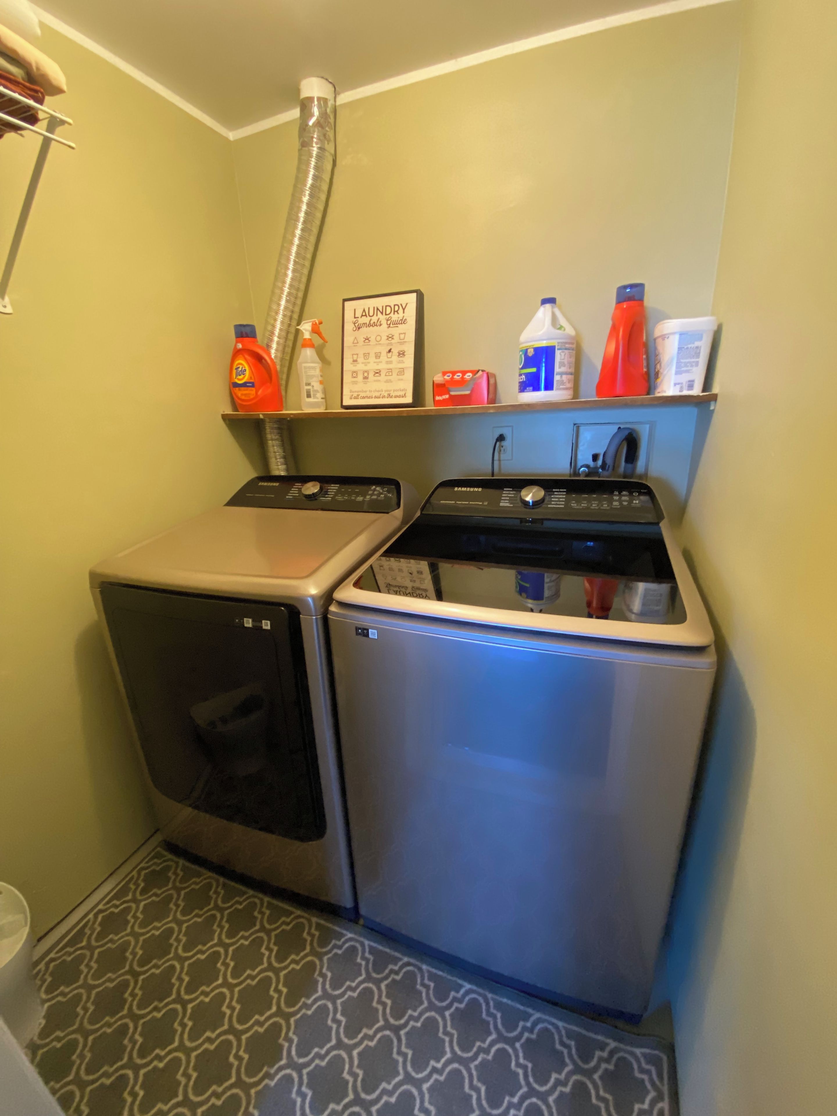 Un espace réduit dédié à la buanderie, comprenant un lave-linge et un sèche-linge, une étagère avec des produits de nettoyage et un guide illustré des symboles utilisés sur les étiquettes des vêtements.