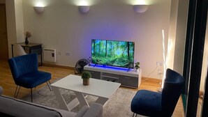 Un salon moderne avec un téléviseur à écran plat, deux fauteuils bleus, une table basse blanche et une petite plante.