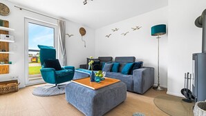 A modern living room with a grey sofa, teal armchair, and a wooden coffee table. There is a large window with curtains, a floor lamp, and wall art. - Lovely apartment in Olpenitz with sauna (Olpenitz)