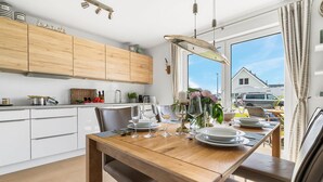 A modern kitchen with wooden cabinets, a dining table set for four, and a view of a house through a large window. - Lovely apartment in Olpenitz with sauna (Olpenitz)