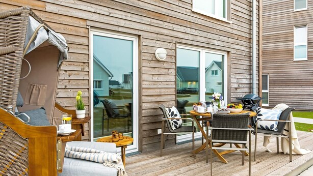 A wooden deck with outdoor furniture, including chairs and a table, and a sliding glass door leading to a patio area. - Lovely apartment in Olpenitz with sauna (Olpenitz)