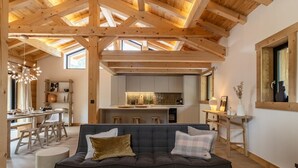 A modern living room with a dark grey sofa, wooden ceiling beams, and a kitchen in the background. - MAISON ORIGINE - BEAUFORT (BEAUFORT)