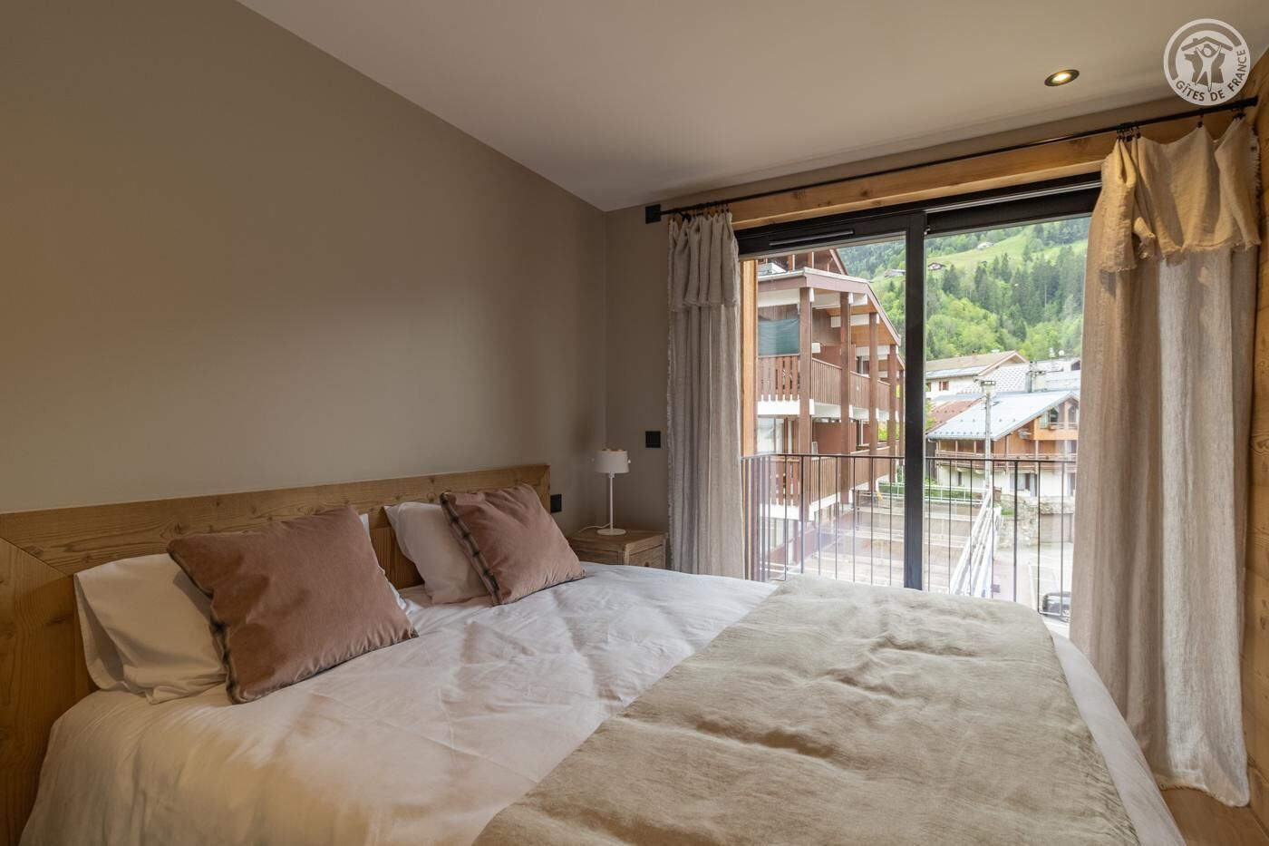 Ein Schlafzimmer mit einem großen Bett, einem Holzkopfende und Blick auf einen Balkon mit Gebäuden und Grünpflanzen.