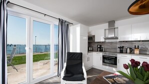 A modern kitchen with white cabinets, a built-in oven, and a coffee maker. There is a dining table with a magazine and a vase of flowers. A chair is placed near a sliding glass door leading to a balcony with a view of the water. - Beautiful apartment in Olpenitz (Olpenitz)