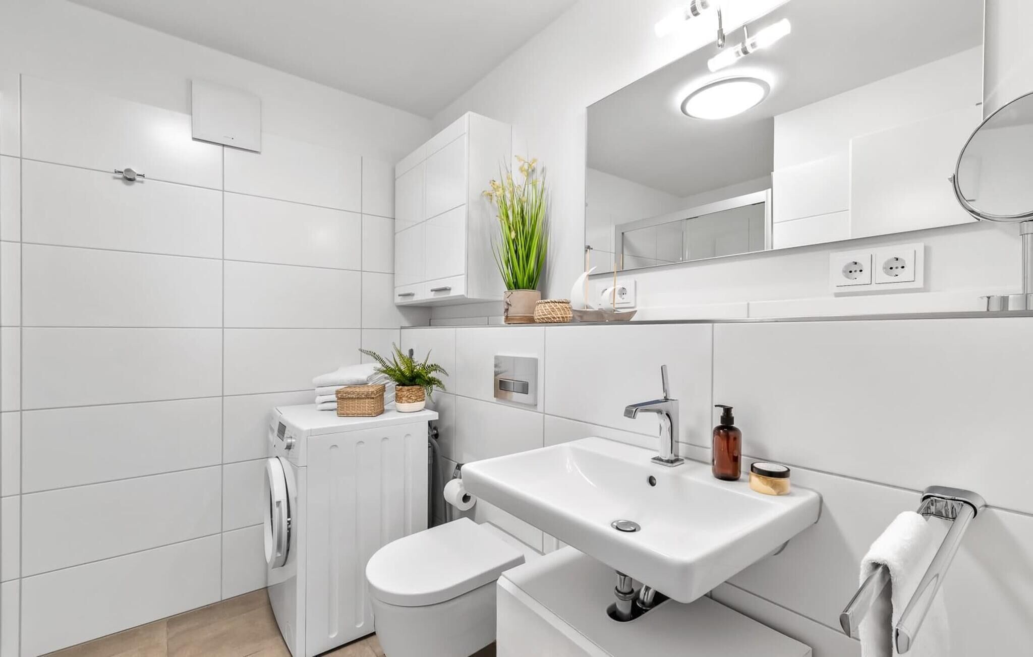 Un baño moderno con lavamanos, inodoro y lavadora blancos. Hay plantas y artículos de aseo en la encimera.