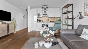 Un salon moderne avec un canapé gris, une table basse agrémentée d’objets décoratifs, un téléviseur et un coin cuisine doté de meubles blancs et d’une chaise de salle à manger.