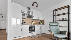 Une cuisine moderne avec des meubles blancs, un four encastré et une machine à café. On y trouve une chaise verte et une étagère en bois ornée d’objets décoratifs.