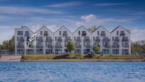 Un immeuble résidentiel moderne à plusieurs étages, doté de balcons, situé près d’un plan d’eau.