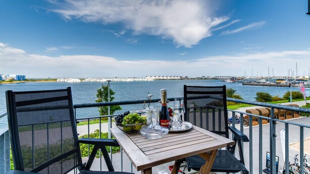 Une table dressée avec une bouteille de champagne, un verre et un saladier de fruits, placée sur un balcon donnant sur une marina, avec des bateaux et des bâtiments en arrière-plan.