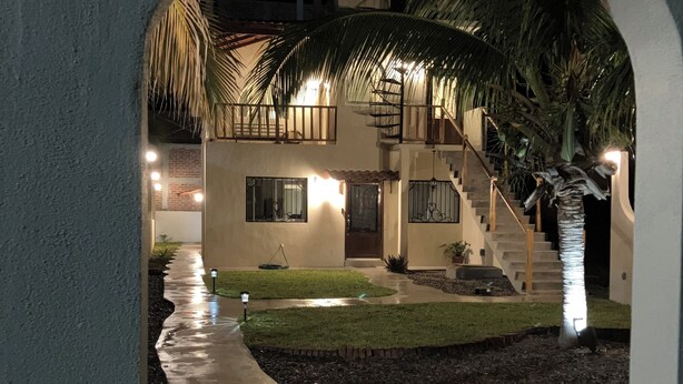 A nighttime view of a building with a covered walkway, palm trees, and a staircase leading to an upper level. - Los Amores Apartments C (Perula)