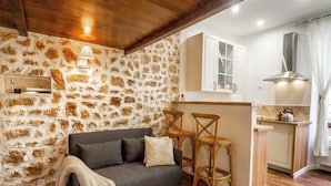Ein gemütlicher Wohnbereich mit Steinwand, grauem Sofa, Holzstühlen und einer Küche mit weißen Schränken und einer Edelstahl-Dunstabzugshaube.