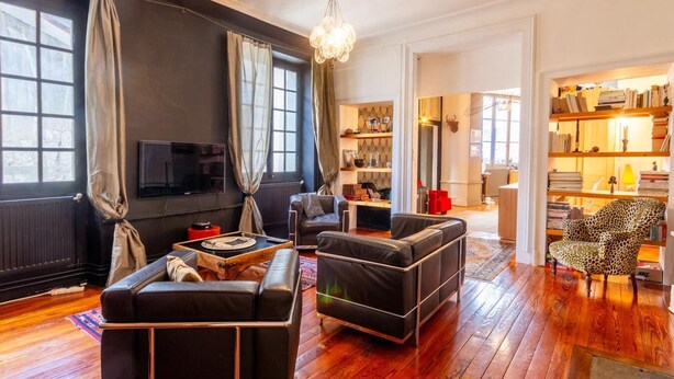 Un salon moderne avec des canapés en cuir noir, une table basse en bois et un téléviseur. On y trouve également des étagères remplies de livres et d’objets décoratifs.