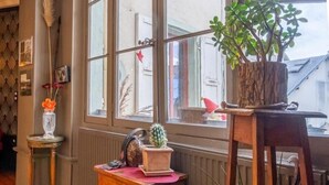 Une pièce chaleureuse avec un plancher en bois, une fenêtre donnant sur des immeubles, une petite table en bois ornée d’une plante et un vase décoratif rempli de fleurs.