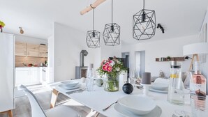 Een moderne eetruimte met een lange witte tafel, geometrische hanglampen en een bloevas.