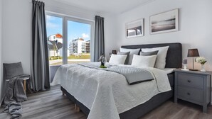 Een moderne slaapkamer met een groot bed, grijze gordijnen, een nachtkastje met een lamp en twee ingelijste foto's aan de muur.