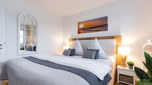 Habitación de hotel moderna con una cama grande, una mesita de noche con lámpara, un espejo y un cuadro de atardecer en la pared.
