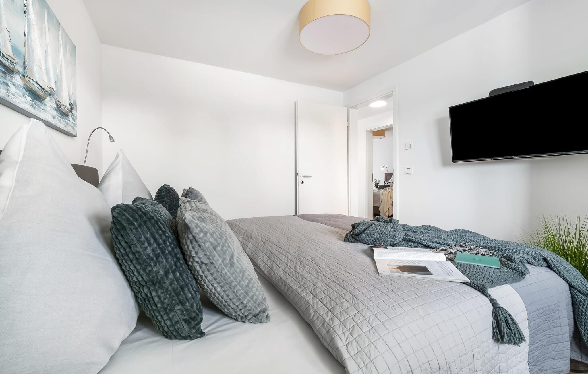 A modern bedroom with a bed, pillows, a bedside table, a lamp, a TV, and a door leading to another room.