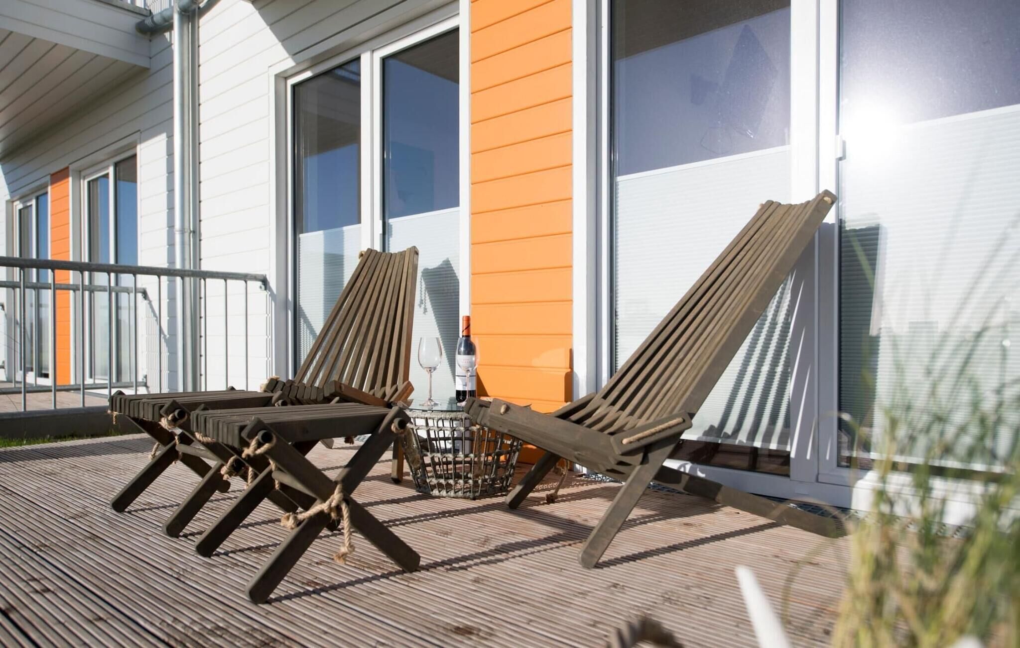 Two wooden lounge chairs on a wooden deck with a glass balustrade, in front of a building with white and orange siding.