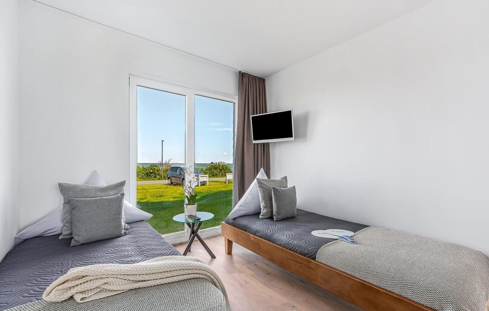 Ein modernes Schlafzimmer mit großem Fenster, Flachbildfernseher und zwei Betten mit grauer Bettwäsche.