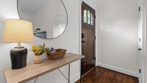 A hallway with a wooden floor, a round mirror, a lamp, a small table with a basket, and a door with a window. - 1 Mile to DT, King Bed, Fenced Yard, Fire Pit, Pet Friendly (Raleigh)