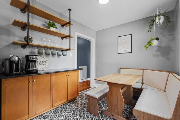 A compact kitchen with wooden cabinets, a coffee machine, and a bench with a cushioned seat. - 1 Mile to DT, King Bed, Fenced Yard, Fire Pit, Pet Friendly (Raleigh)
