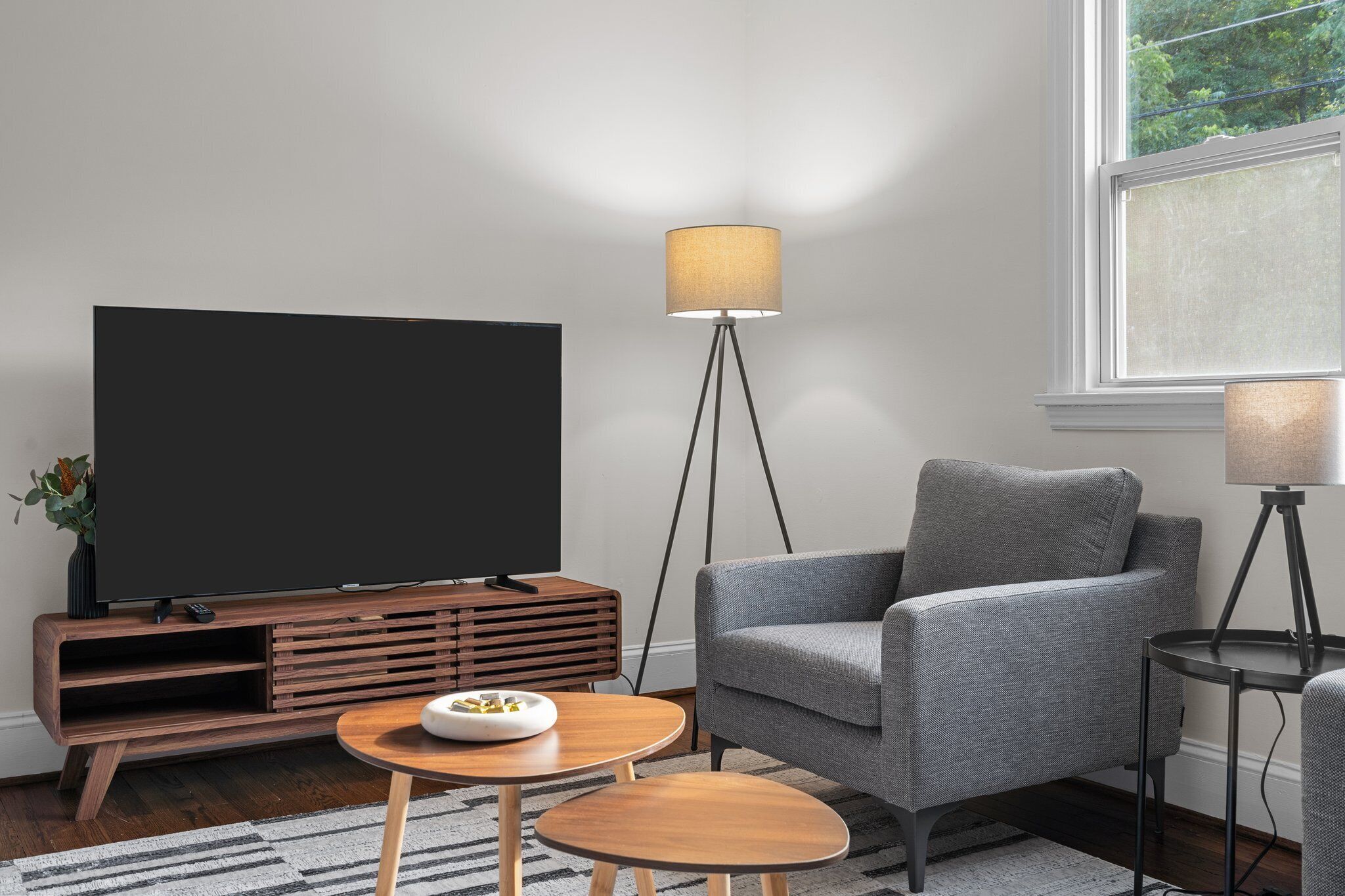 A modern living room with a flat-screen TV, a grey armchair, a round wooden coffee table, and a floor lamp.