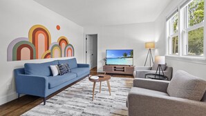 A modern living room with a blue sofa, grey armchair, and a TV on a wooden stand. - 1 Mile to DT, King Bed, Fenced Yard, Fire Pit, Pet Friendly (Raleigh)