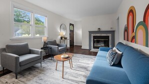 A living room with a fireplace, a blue sofa, a grey armchair, and a small wooden table. - 1 Mile to DT, King Bed, Fenced Yard, Fire Pit, Pet Friendly (Raleigh)