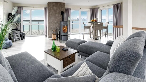 Ett modernt vardagsrum med grå soffor, en centralt placerad öppen spis och en matplats med utsikt över havet.
