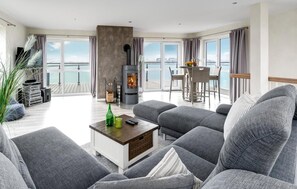 A modern living room with grey sofas, a central fireplace, and a dining area with a view of the sea. - Traumblick auf der Ostsee (Olpenitz)