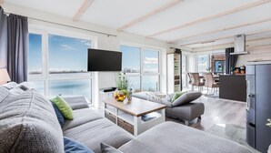 Ett modernt vardagsrum med en stor soffa, ett soffbord och en tv. I anslutning till vardagsrummet finns ett köksområde med ett matbord och stolar, som syns genom en öppen dörr.