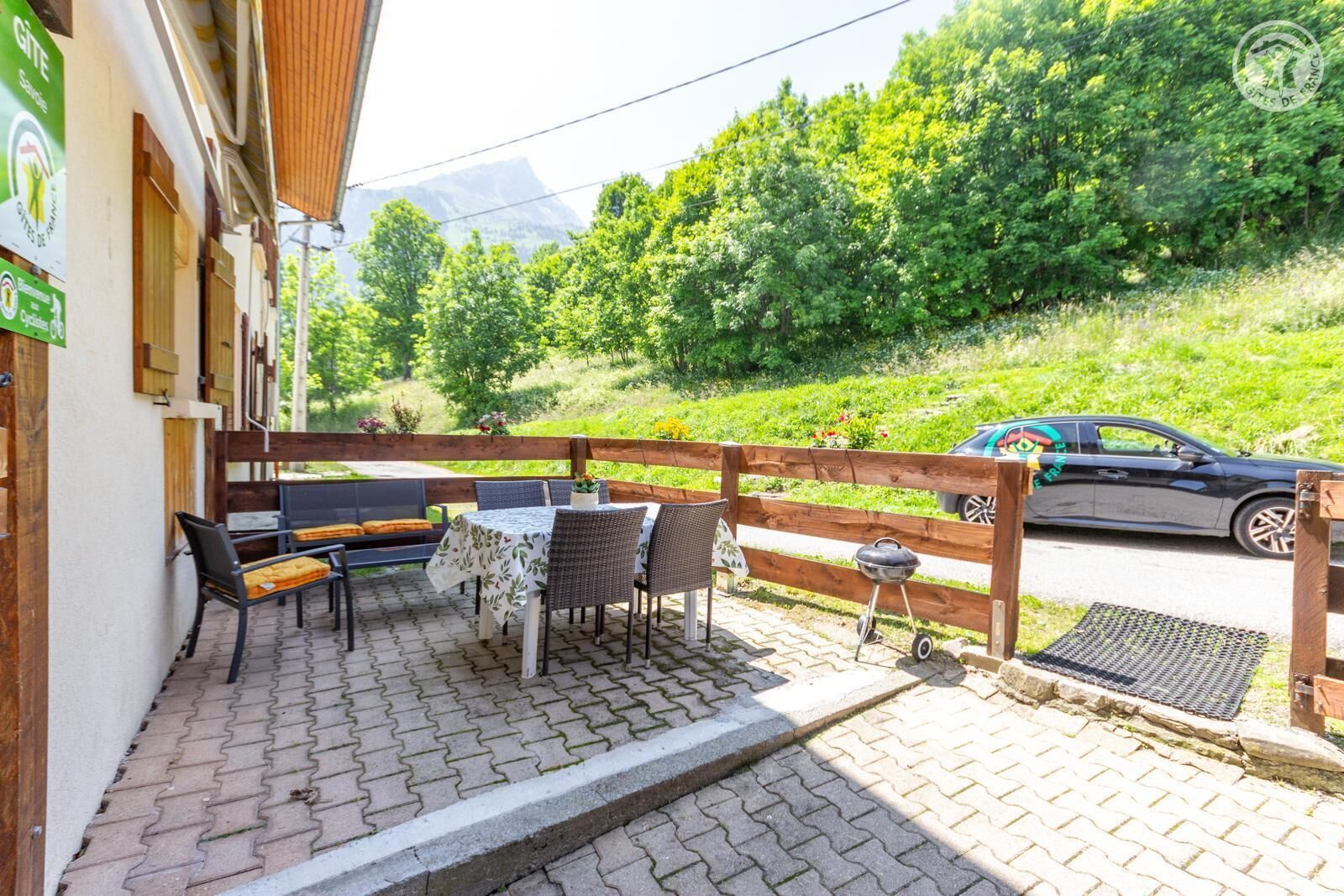 A patio with outdoor seating, a barbecue grill, and a view of a car on a road with greenery in the background.