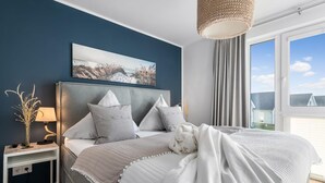 A bedroom with a large bed, a bedside table with a lamp, a painting on the wall, and a view of the outside through a window. - Stunning apartment in Olpenitz (Olpenitz)