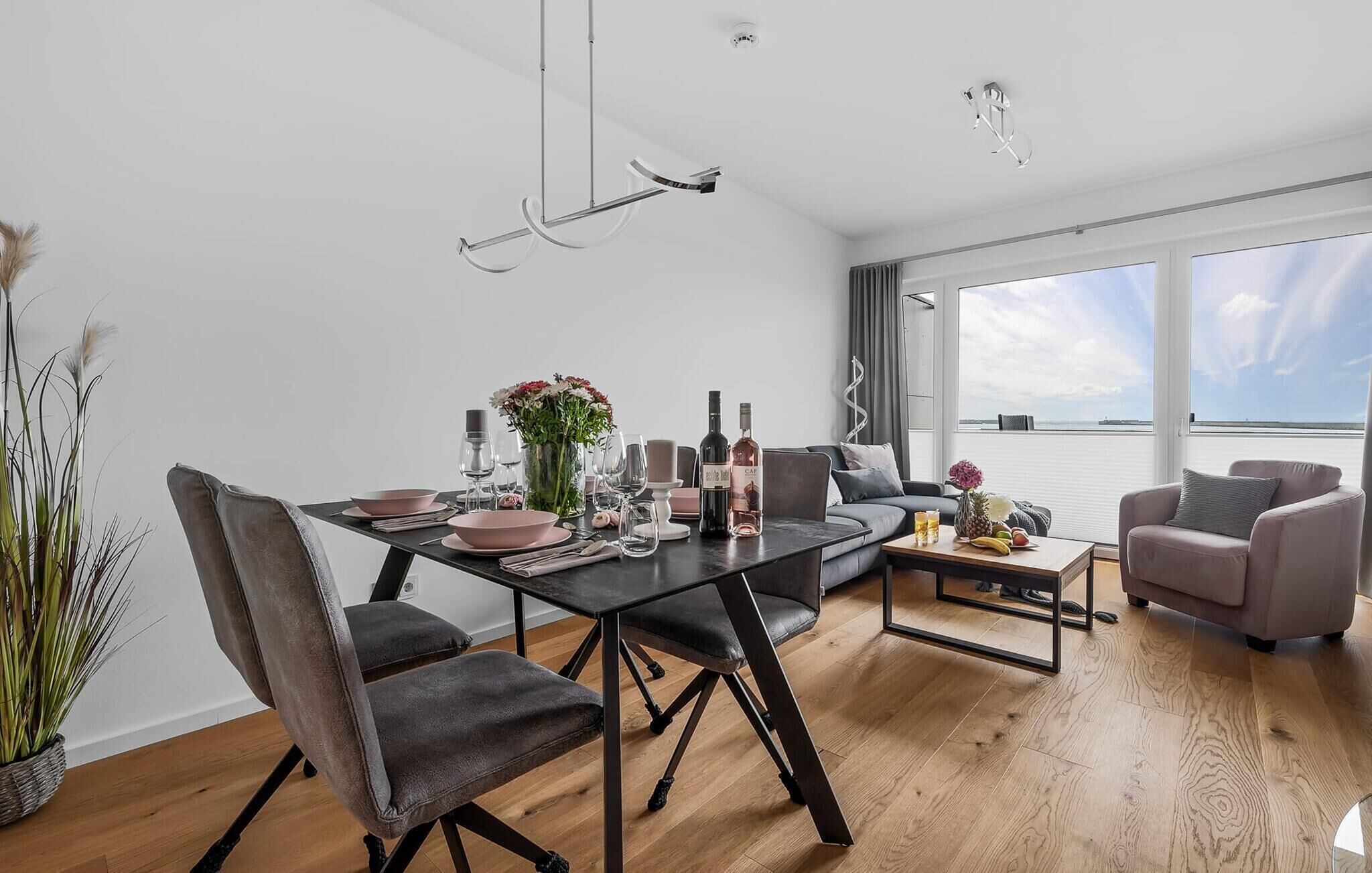 Ein modernes Wohnzimmer mit Essbereich, ausgestattet mit einer Couch, Sesseln, einem Couchtisch und einem Esstisch, der mit Gläsern und Blumen gedeckt ist.