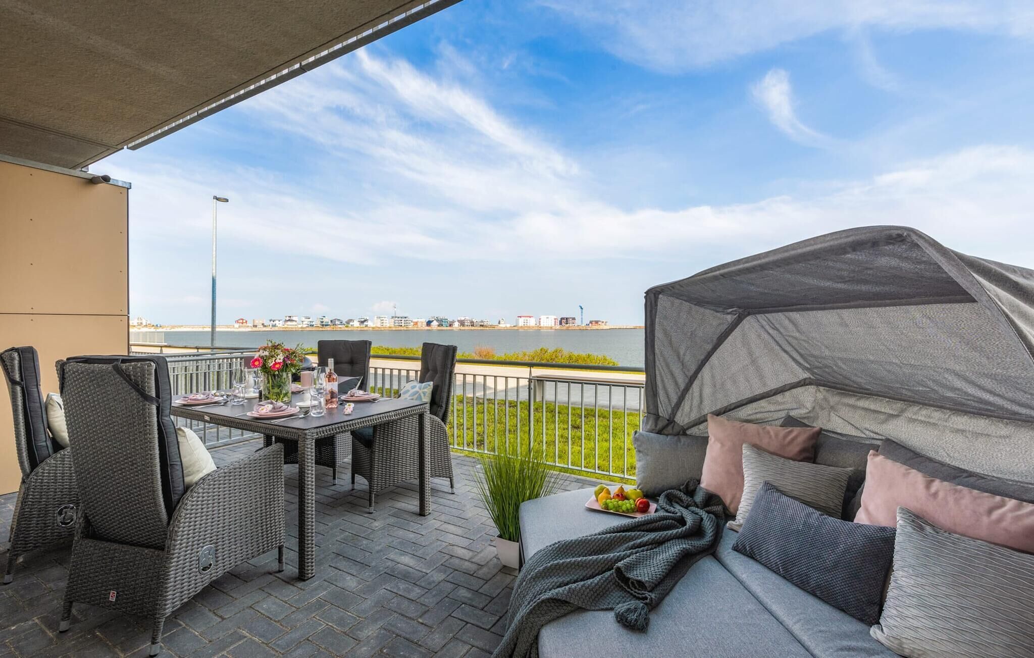 Een terras buiten met rieten meubilair, een eettafel en een overdekt zitgedeelte met kussens.