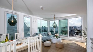 Uma sala de estar moderna com sofá, mesa de centro e área de jantar. Janelas amplas oferecem vista para a cidade.