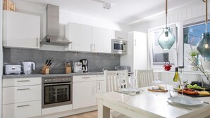 Cozinha moderna com armários brancos, eletrodomésticos de aço inoxidável e área de refeições com mesa e cadeiras brancas.