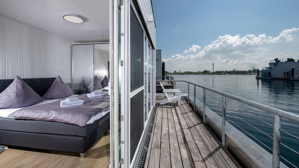 Quarto moderno com cama, vista para a água e varanda com guarda-corpo.