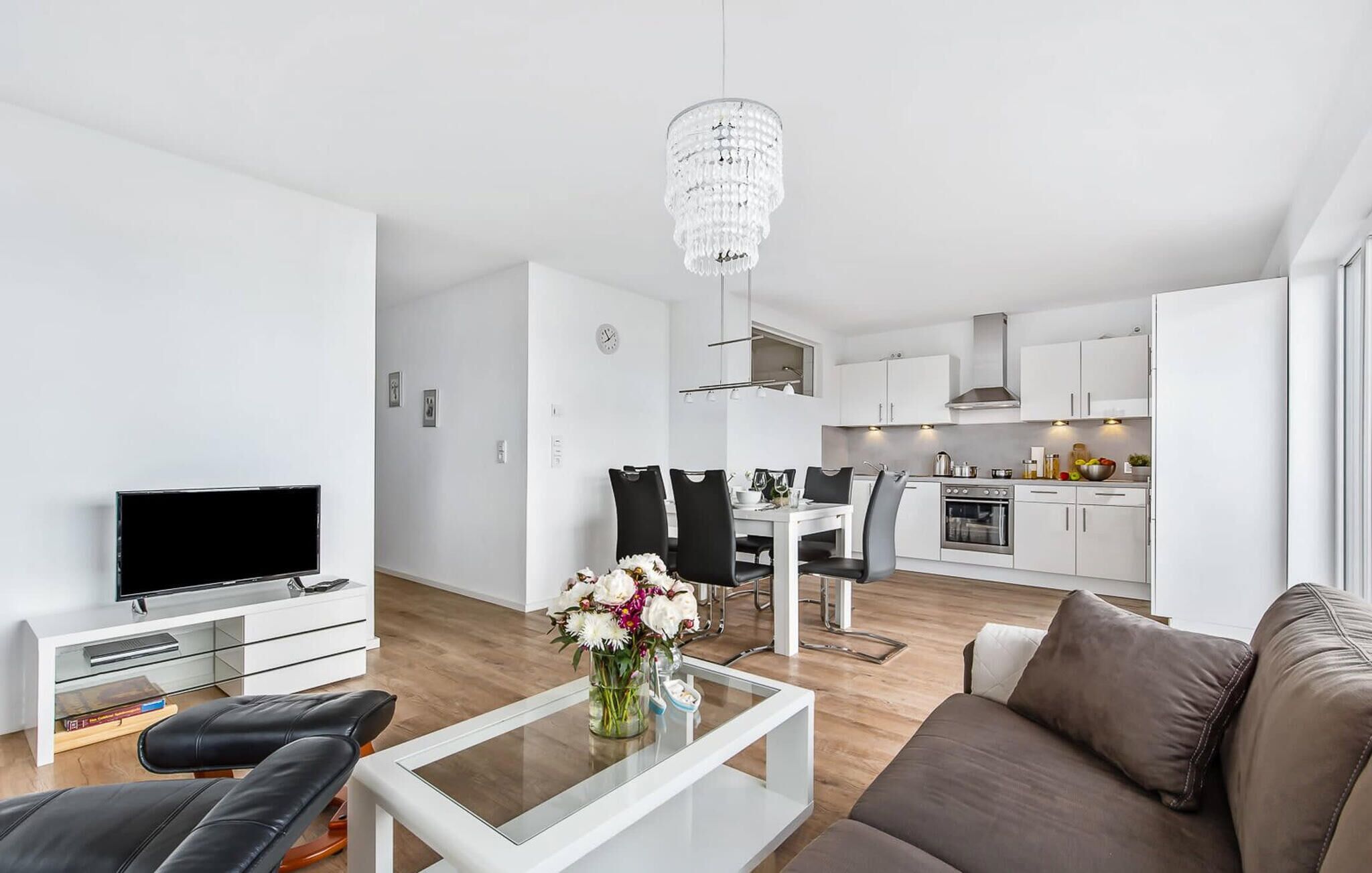 Un salon moderne avec un téléviseur à écran plat, une table basse en verre ornée d’un vase de fleurs, un canapé marron et un coin repas avec une table et des chaises blanches.