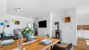 Un salon moderne avec une table à manger en bois, préparée pour quatre personnes, une cheminée, un téléviseur à écran plat et un coin salon confortable agrémenté d’un fauteuil à motifs.