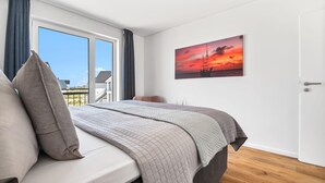 Une chambre avec un grand lit, une vue sur la mer par une porte coulissante et un tableau représentant un coucher de soleil accroché au mur.