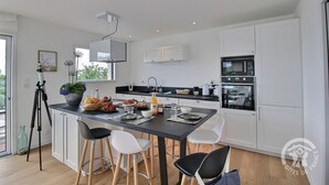 A modern kitchen with white cabinets, a black countertop, and stainless steel appliances. There is a dining area with a table set for two, and a telescope on a tripod in the corner. - Le Menhir - PLEUMEUR BODOU (PLEUMEUR BODOU)