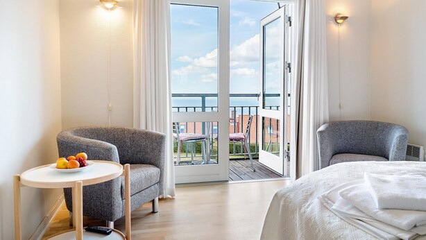 Une chambre d’hôtel avec deux fauteuils gris, une petite table ronde sur laquelle sont disposés des fruits, un lit avec du linge de maison blanc et un balcon offrant une vue sur la mer et les bâtiments.