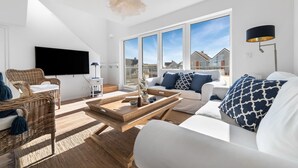 A modern living room with a flat-screen TV, wicker furniture, a white sofa with patterned cushions, and large windows offering a view of neighboring houses.
