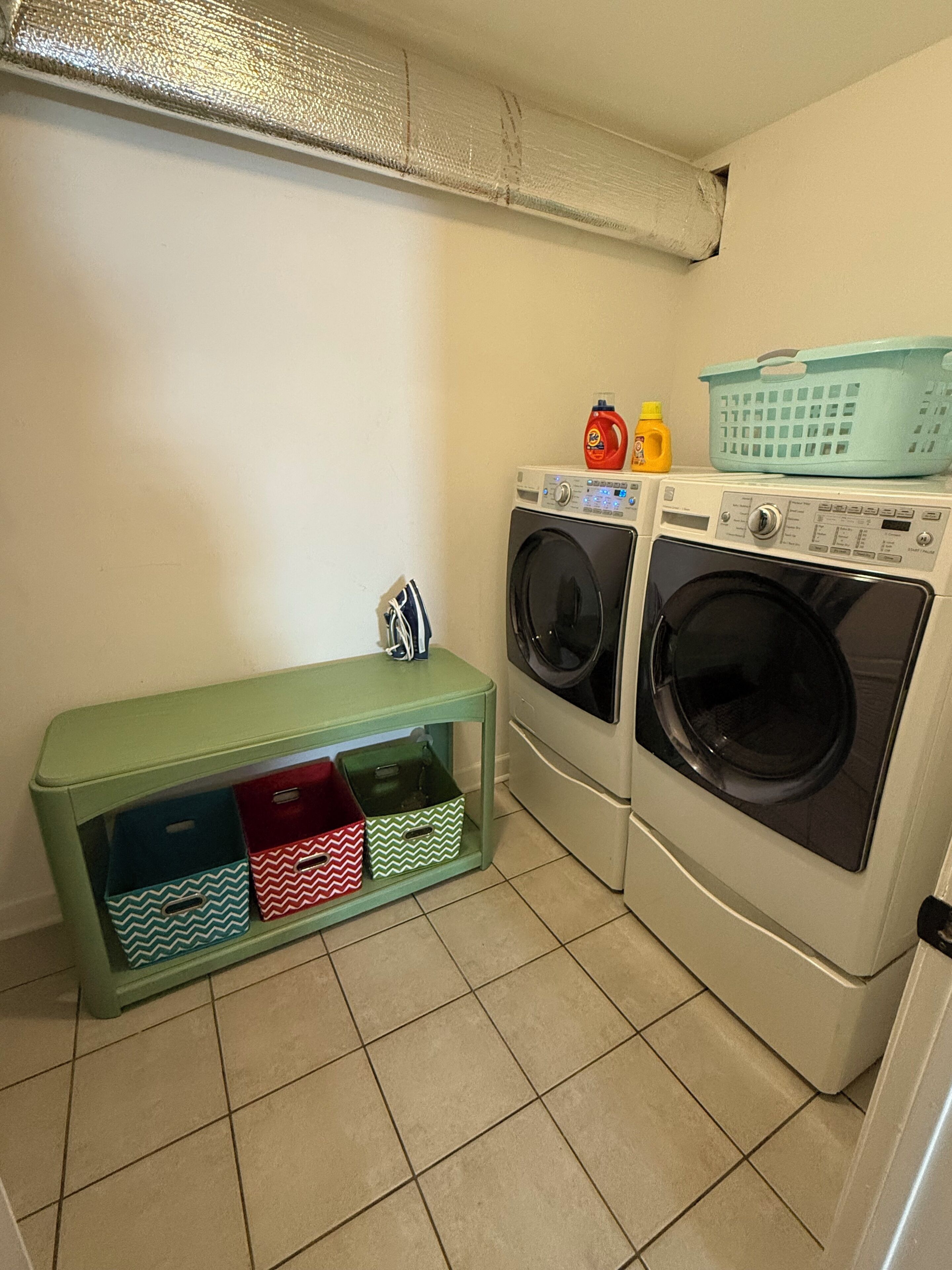 A laundry area with two washing machines, a green storage unit with colorful bins, and a laundry basket on top of the machines.