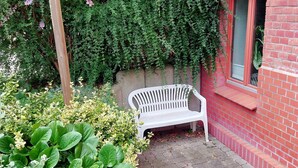 Eine weiße Plastikbank vor einer roten Backsteinmauer, umgeben von Grünpflanzen und einem Backsteinweg.