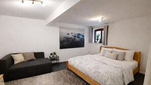 Ein Schlafzimmer mit einem Bett, einem Sofa, einem kleinen Tisch mit Pflanzen, einem Bild an der Wand und einem Fenster mit Vorhängen.