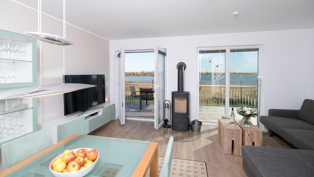 A modern living room with a TV, a dining area with a glass table, a sofa, and a large window overlooking a body of water. - Nice home in Olpenitz with sauna (Olpenitz)