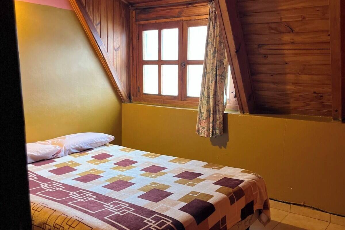 Un dormitorio con techo de madera inclinado, una cama con cubrecama estampado, una ventana con cortinas y una pared amarilla.