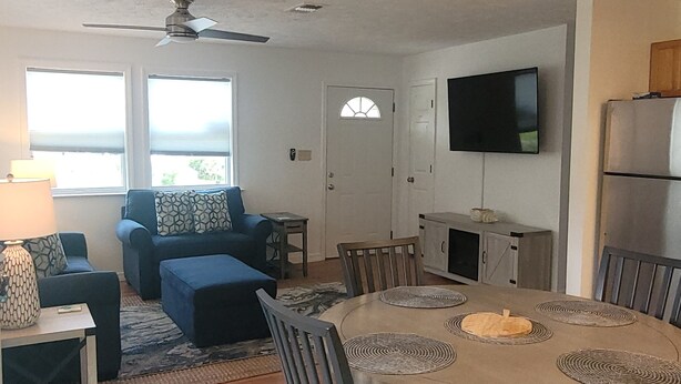 A living room with a sofa, ottoman, and a dining table with chairs. There is a ceiling fan, a TV mounted on the wall, and a refrigerator.